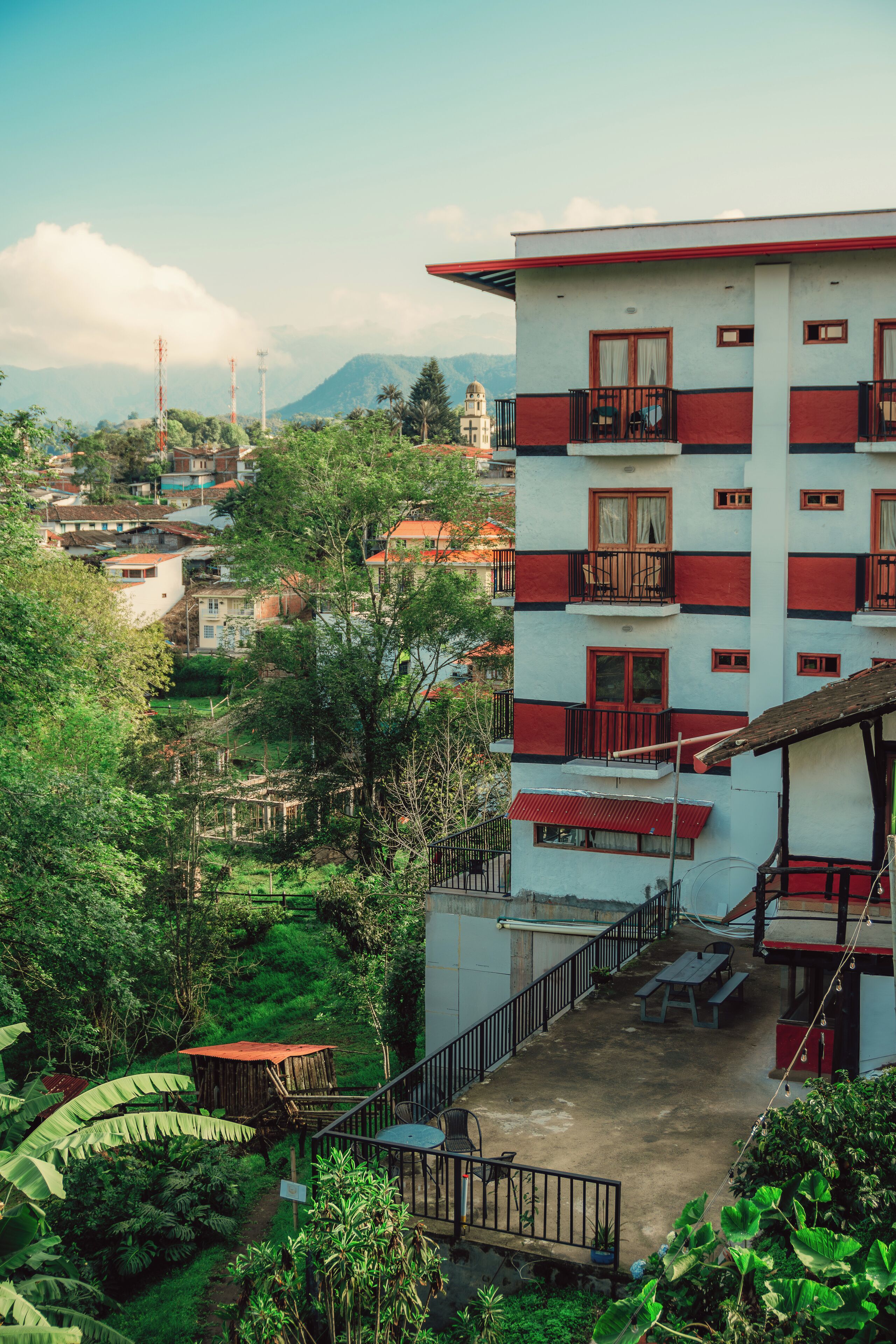 Un edificio de varios pisos con balcones, rodeado de abundante vegetación y montañas al fondo.