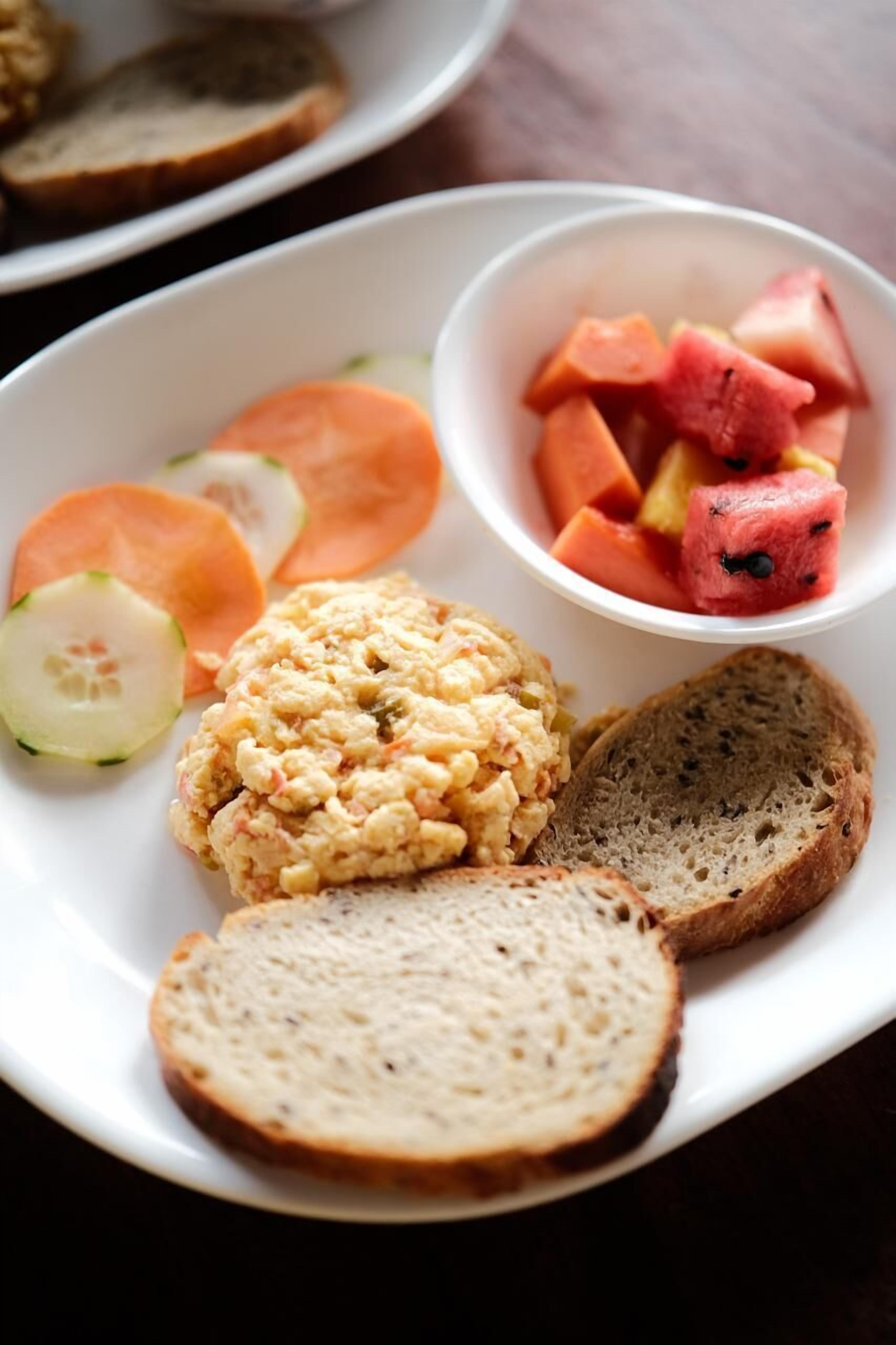 A plate with scrambled eggs, cucumber slices, carrot sticks, watermelon cubes, and two slices of bread.