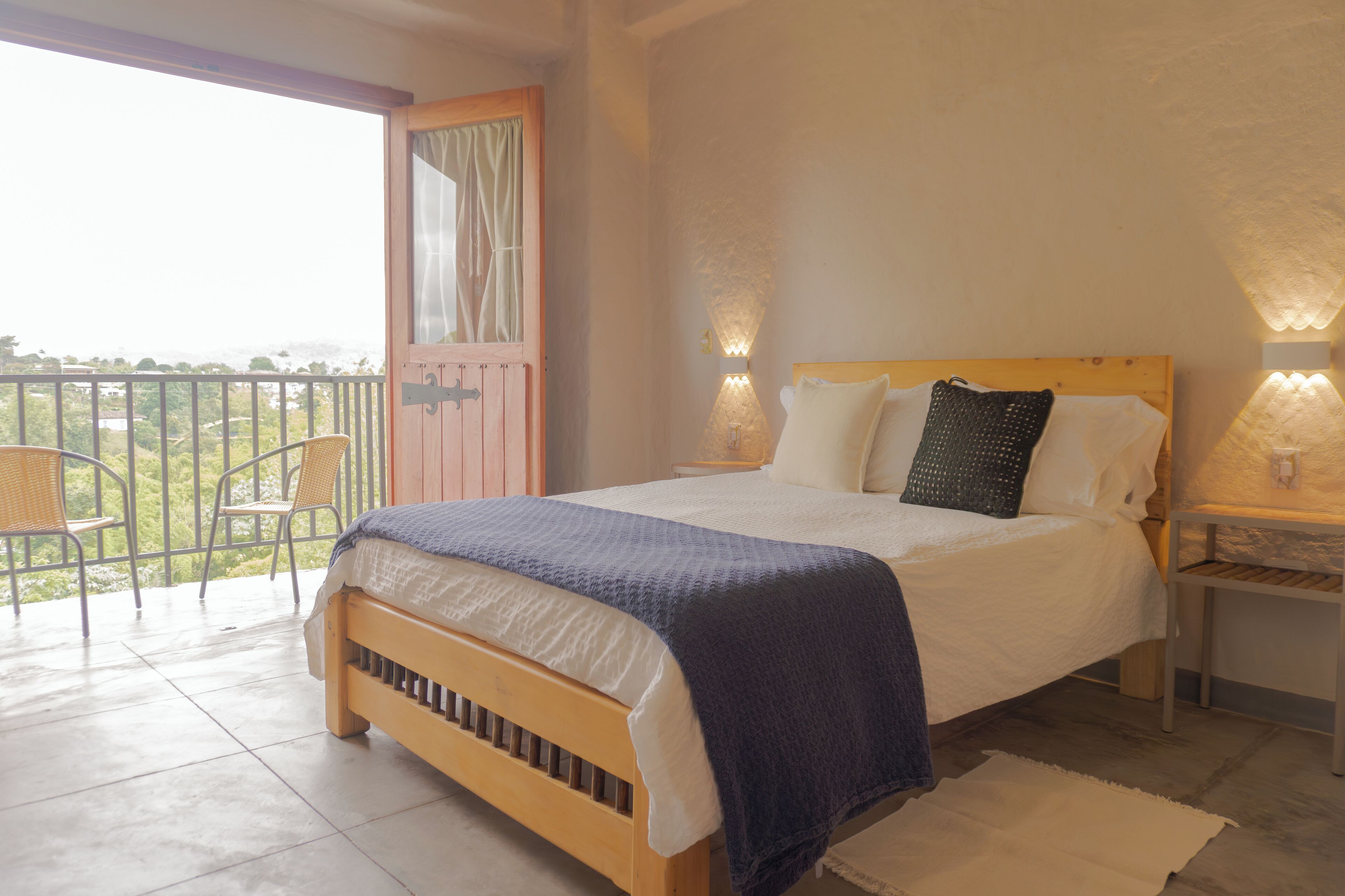 A bedroom with a wooden bed, two bedside tables with lamps, a balcony with chairs, and a view of greenery.
