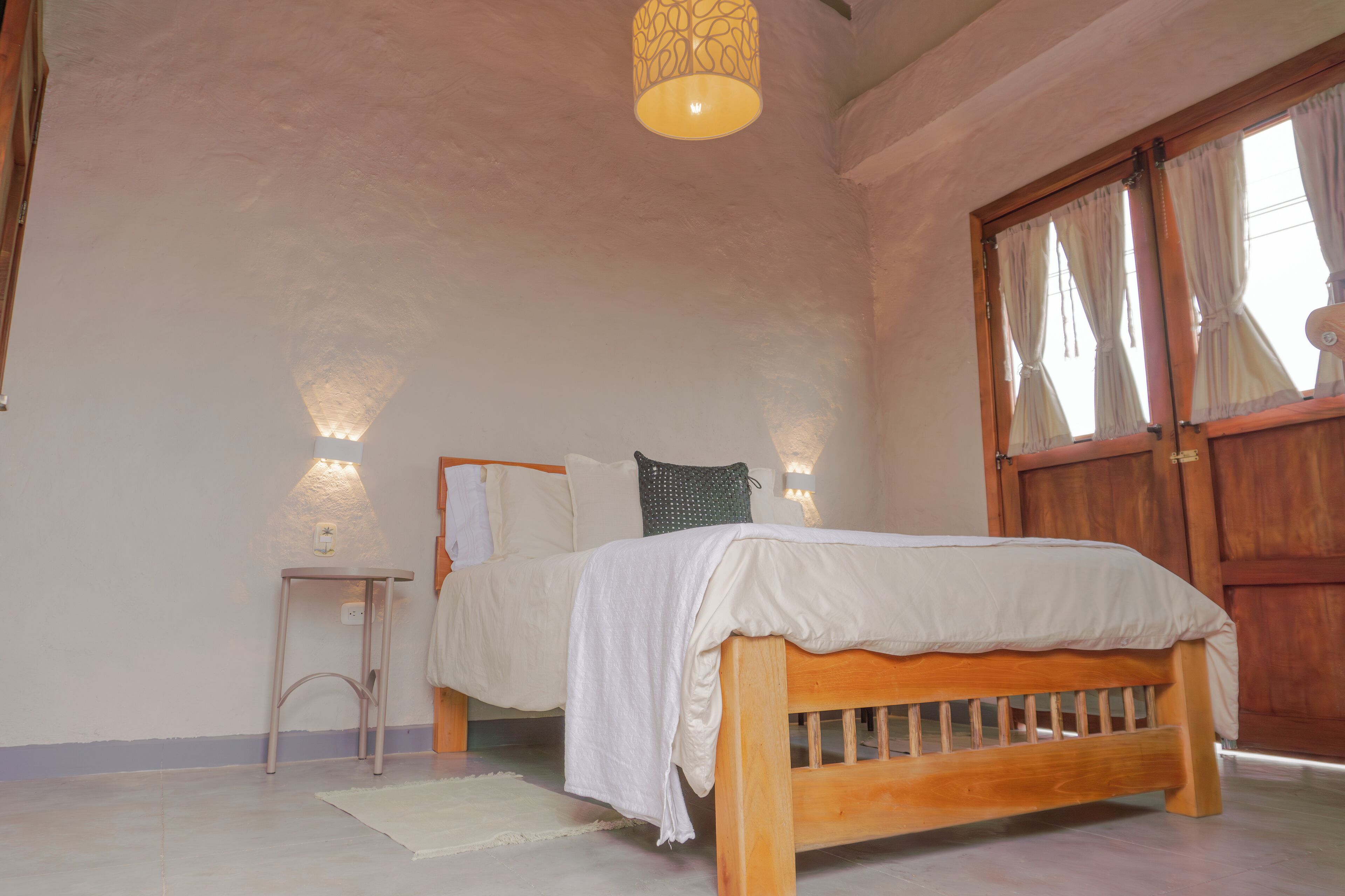 Ein Schlafzimmer mit einem hölzernen Bett, einem Nachttisch, einer Lampe und einem großen Fenster mit Vorhängen.