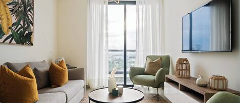 Een moderne woonkamer met een grijze bank, een groene fauteuil en een ronde salontafel. Een groot raam met dunne gordijnen laat natuurlijk licht binnen, en een flatscreen televisie is aan de muur bevestigd.