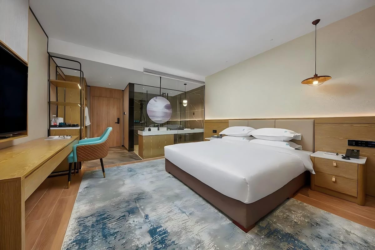 A modern hotel room with a large bed, a desk, a chair, a TV, a wardrobe, and a bathroom visible through a glass partition.