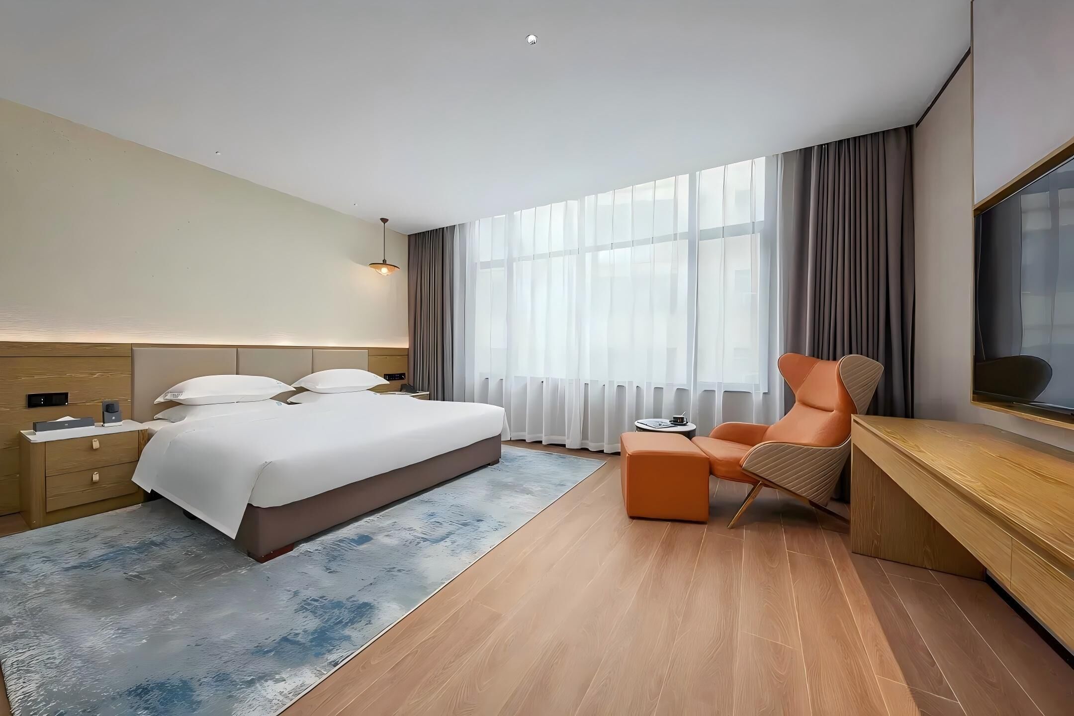 A modern hotel room with a large bed, a side table, a small round table, an orange chair, a wooden desk, and a large window with curtains.