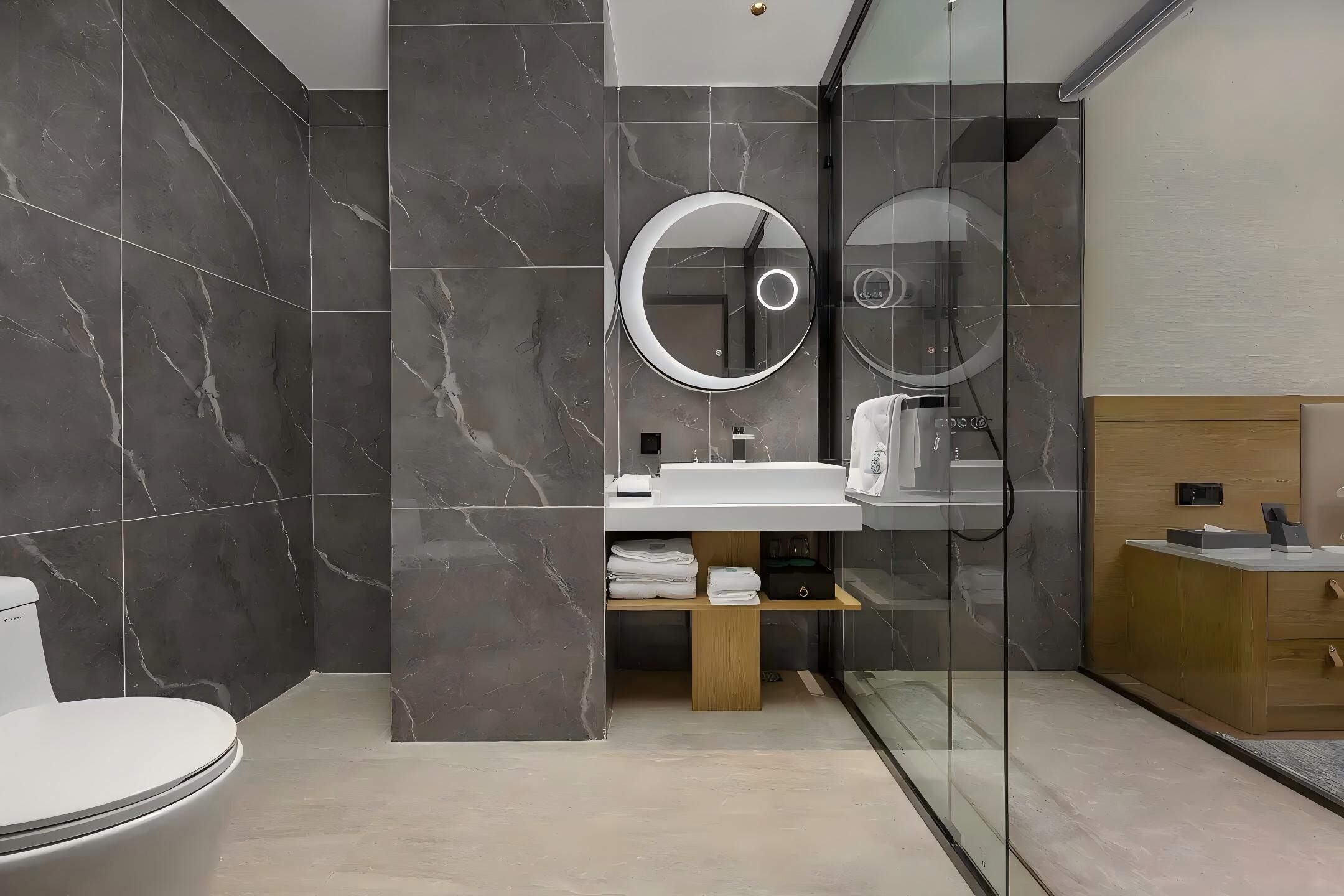 Une salle de bain moderne avec des carreaux de marbre foncé, des toilettes blanches, un lavabo avec un miroir et un meuble-lavabo en bois.