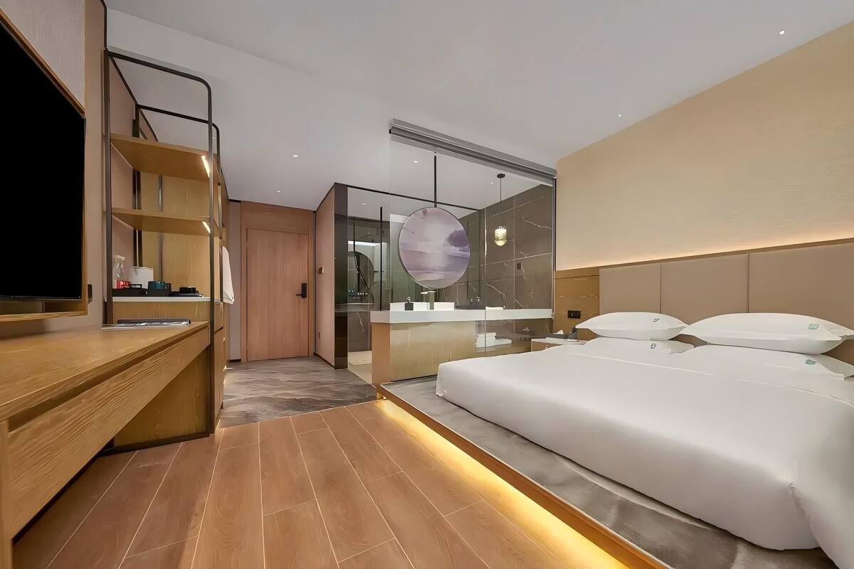 A modern hotel room with a wooden desk, a bed with white bedding, a large mirror, and a bathroom with a glass partition.
