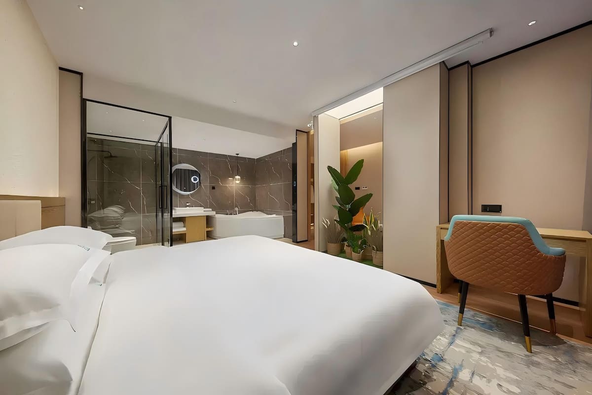 A hotel room with a large bed, a desk, a chair, a bathroom with a bathtub, and a potted plant.