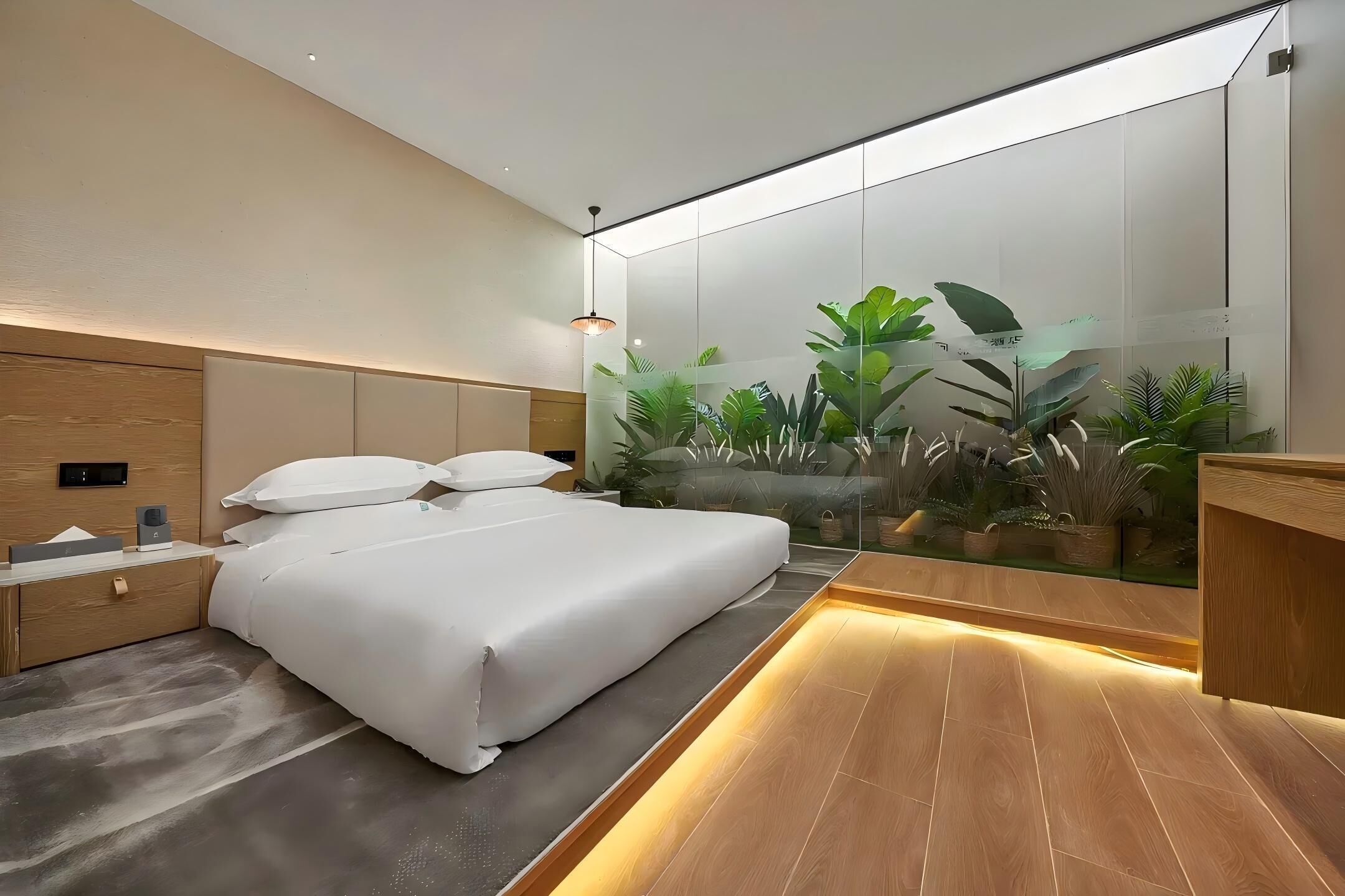 A modern bedroom with a large bed, bedside tables, a nightstand, a wooden headboard, a glass partition with plants, and a built-in desk area.