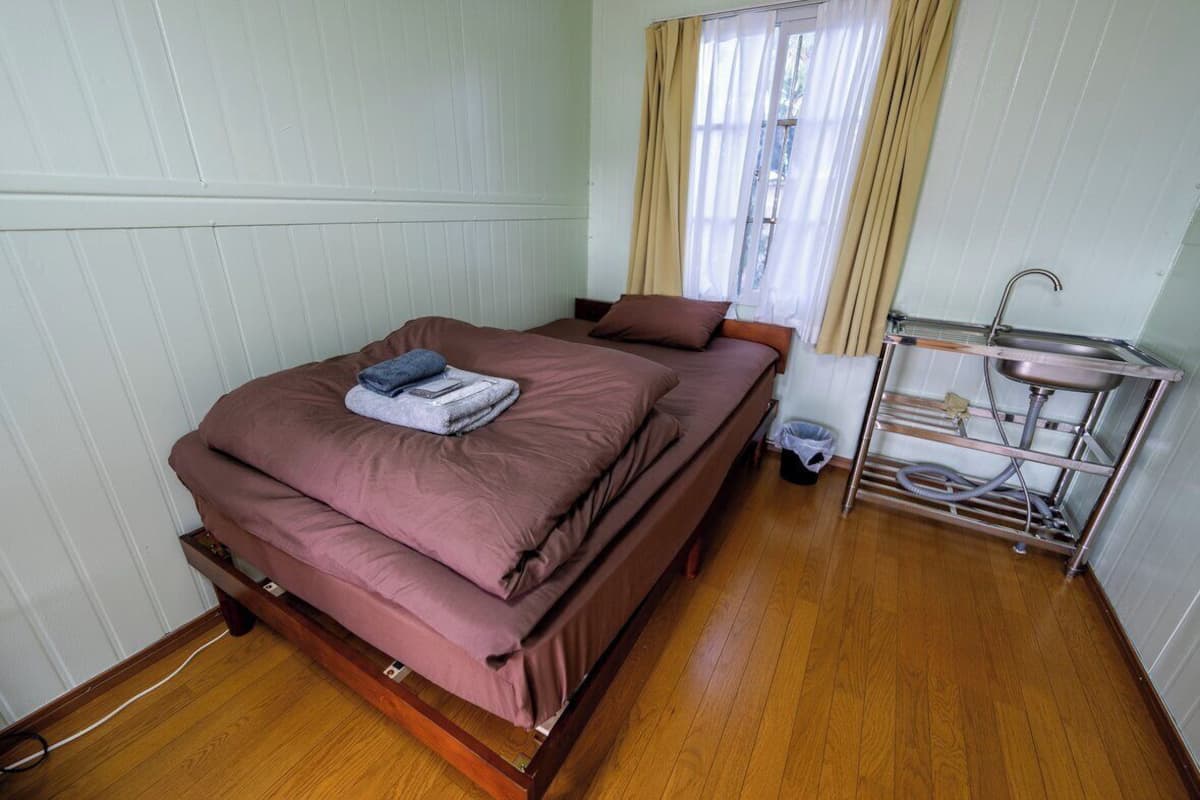 A single bed with a wooden frame, a metal shelf unit with a sink, and a window with curtains.