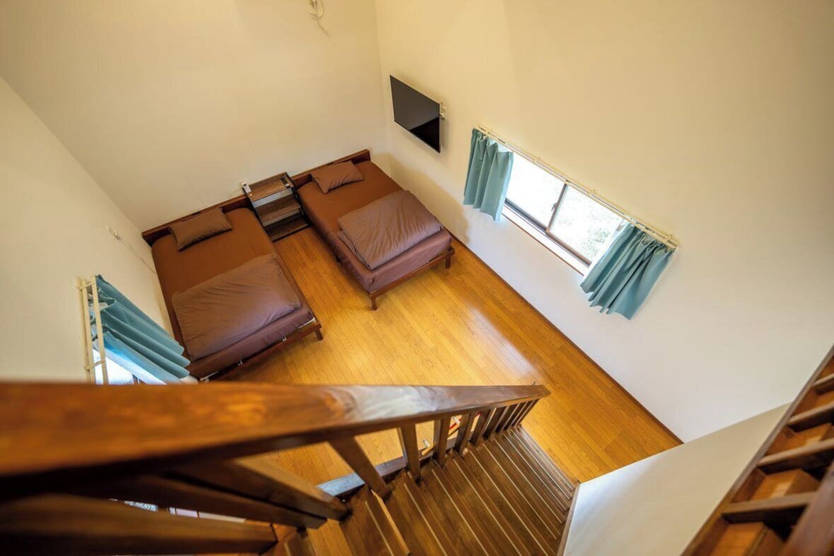 A room with two beds, a TV mounted on the wall, and a window with curtains.