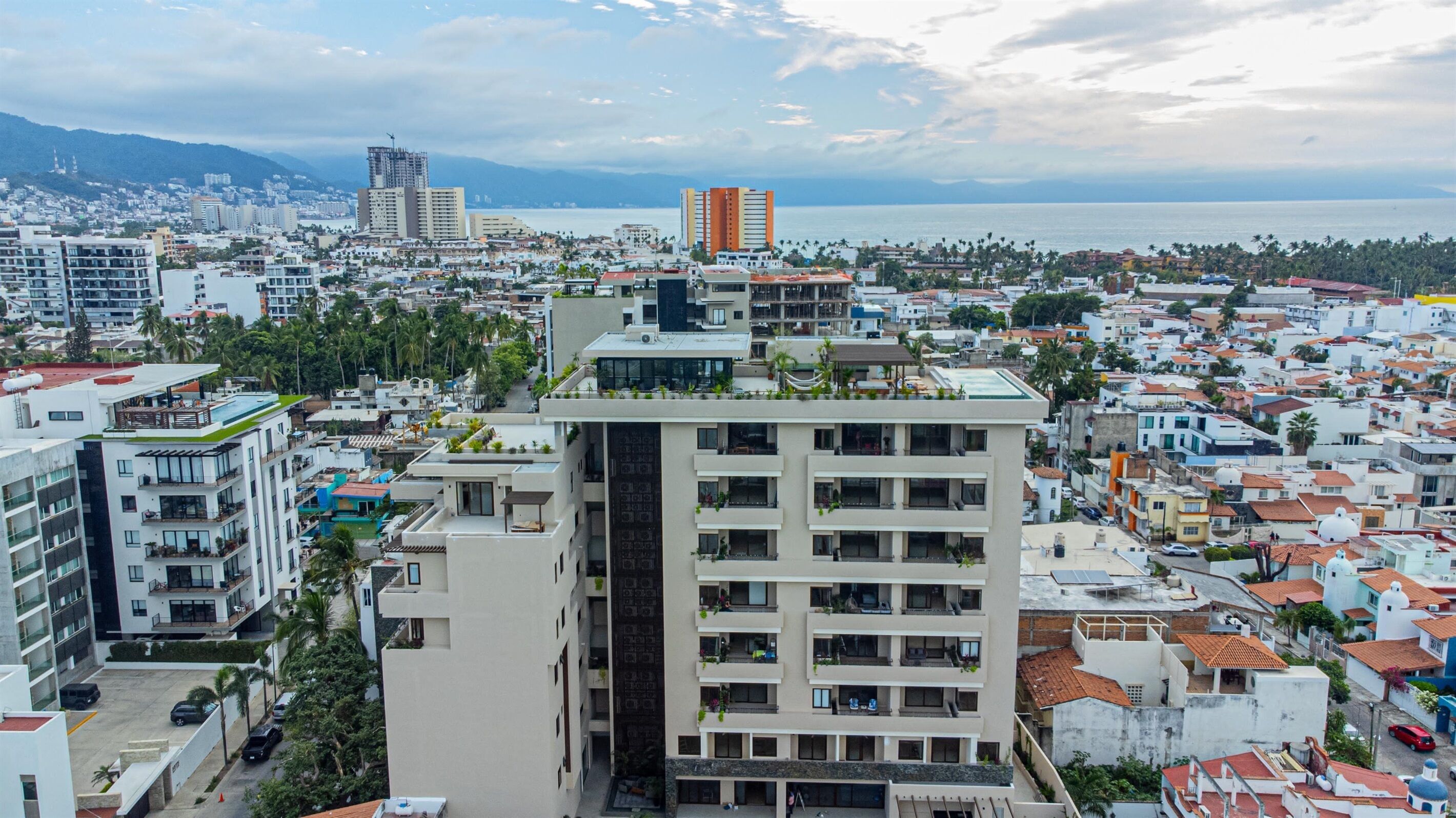 A high-rise building with multiple balconies, surrounded by a densely packed urban area with various buildings and a coastline in the background.