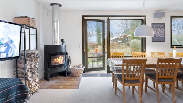 A modern living room with a wood-burning stove, a dining area with wooden chairs, and a large window offering a view of the outdoors. - Lovely home in Samsø with WiFi (Samsø)
