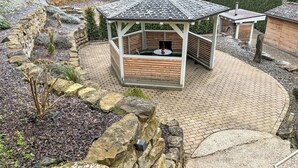 Un belvédère doté d’une terrasse en bois et d’un toit en tuiles, entouré d’un jardin aménagé avec des rochers et des arbustes.