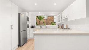 Eine moderne Küche mit weißen Schränken, einem Edelstahl-Kühlschrank, einem eingebauten Backofen und einem großen Fenster mit Blick auf eine Ziegelwand und Grünflächen.
