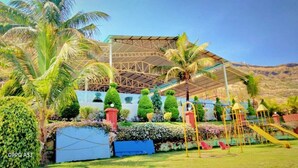 A covered outdoor area with a playground, palm trees, and a building with a metal roof. - Madhura Agro Tourism (Wai)