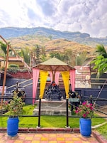 A temple with a statue of a deity under a canopy, surrounded by greenery and a mountainous backdrop.