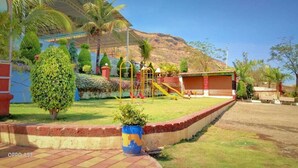 A playground with slides and climbing structures, surrounded by well-maintained grass and palm trees, with a mountain in the background. - Madhura Agro Tourism (Wai)
