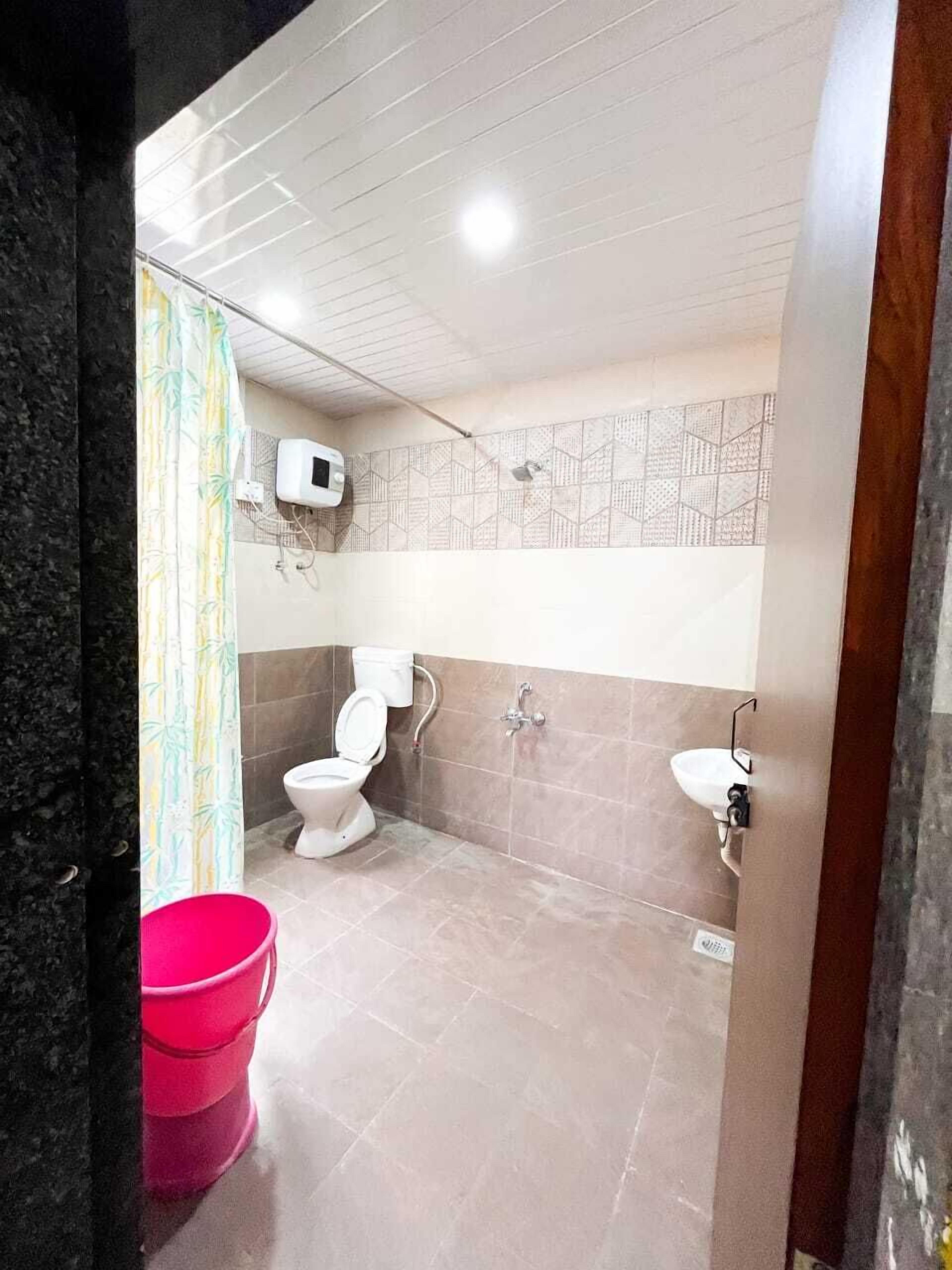 Casa de banho com sanita, lavatório e chuveiro. Há um balde rosa e uma cortina de chuveiro estampada.
