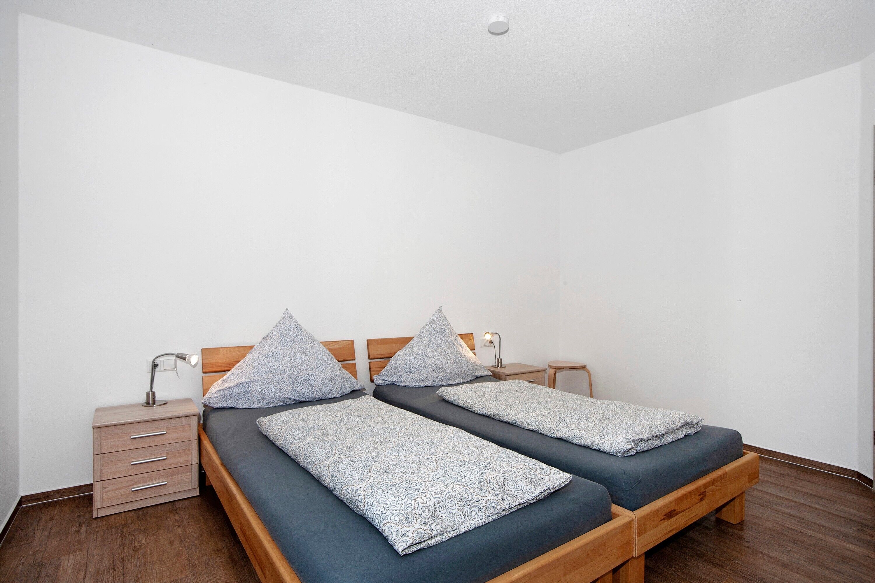 A room with two single beds, a nightstand, a lamp, and a wooden floor.