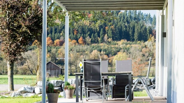 A covered outdoor area with a table and chairs, a metal railing, and a view of a forested hillside. - Apartment 'Im Allgäu - Biber 1' with Private Terrace, Shared Garden and Wi-Fi (Leutkirch im Allgäu)