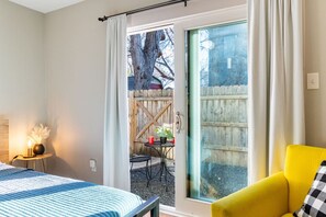 A bedroom with a bed, a yellow chair, a small table with a plant, and a sliding glass door leading to an outdoor area with a table and chairs. - Huge 5BR compound! Walk to downtown & Rainey St. (Austin)