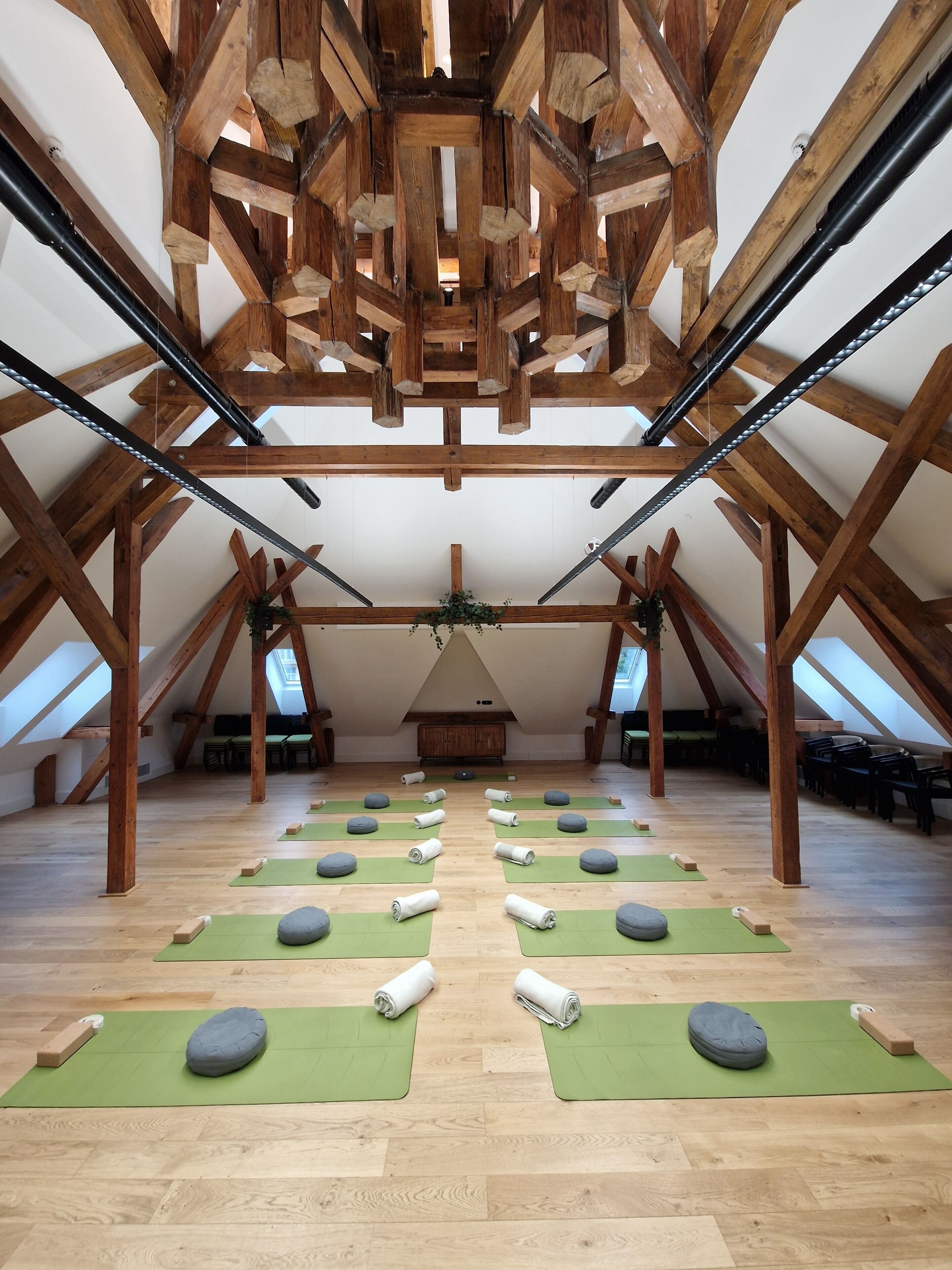 Una stanza spaziosa con soffitto alto sostenuto da travi in legno, e file di tappetini da yoga verdi su pavimento in legno.