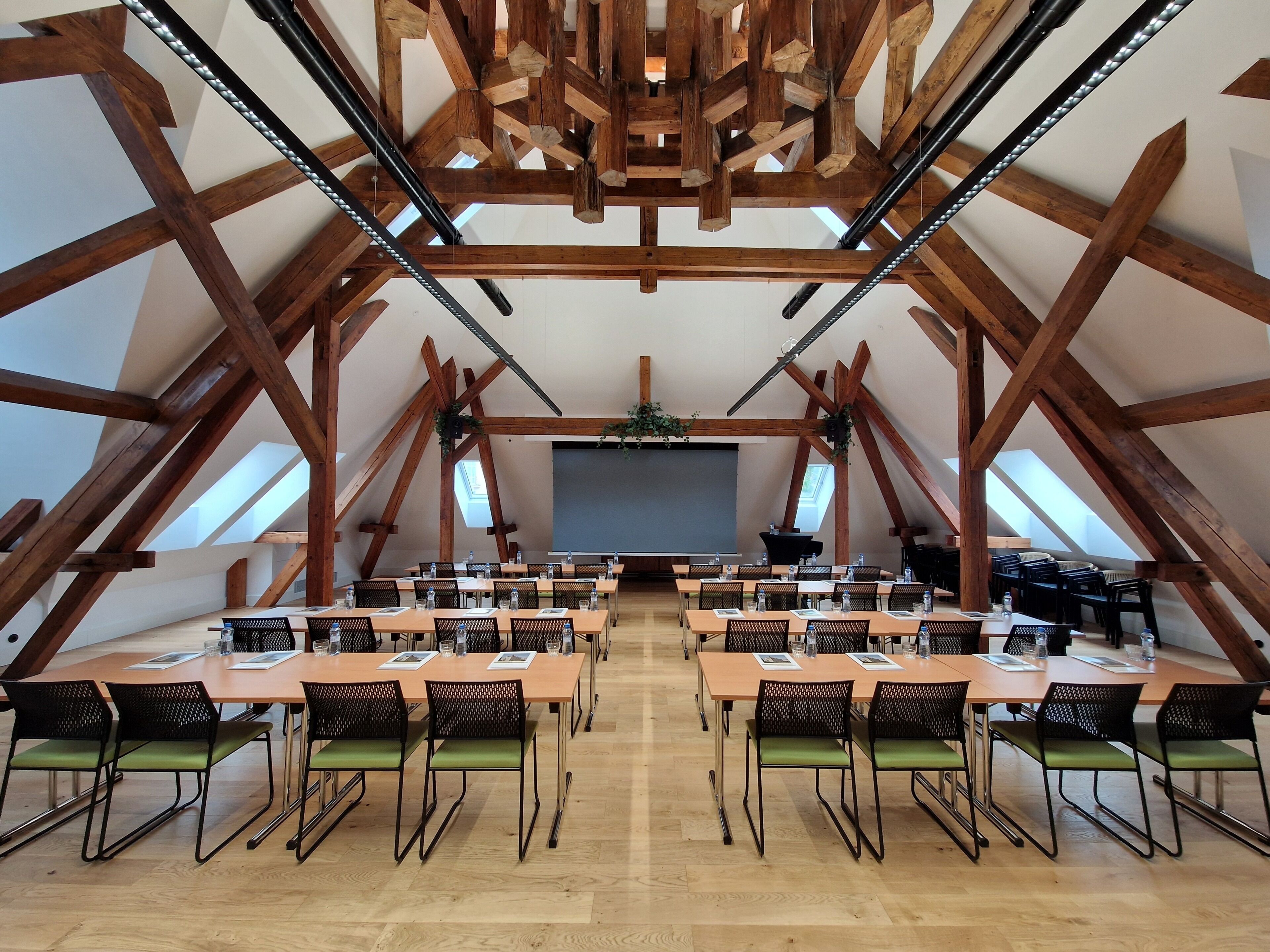 A large, open-plan room with high wooden beams, multiple tables with chairs, and a projector screen at the front.