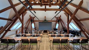 A large, open-plan room with high wooden beams, multiple tables with chairs, and a projector screen at the front.