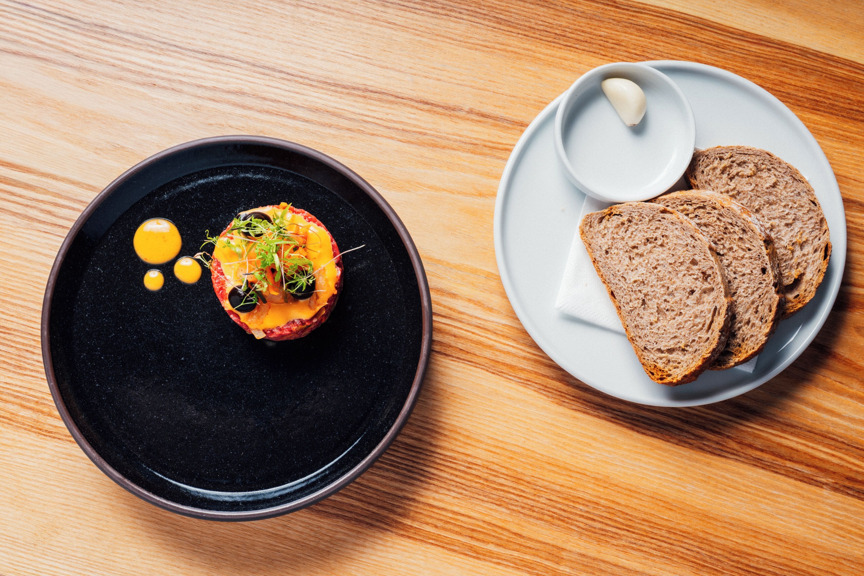 A table with a black plate containing a food item and a white plate with slices of bread and a small bowl of butter.