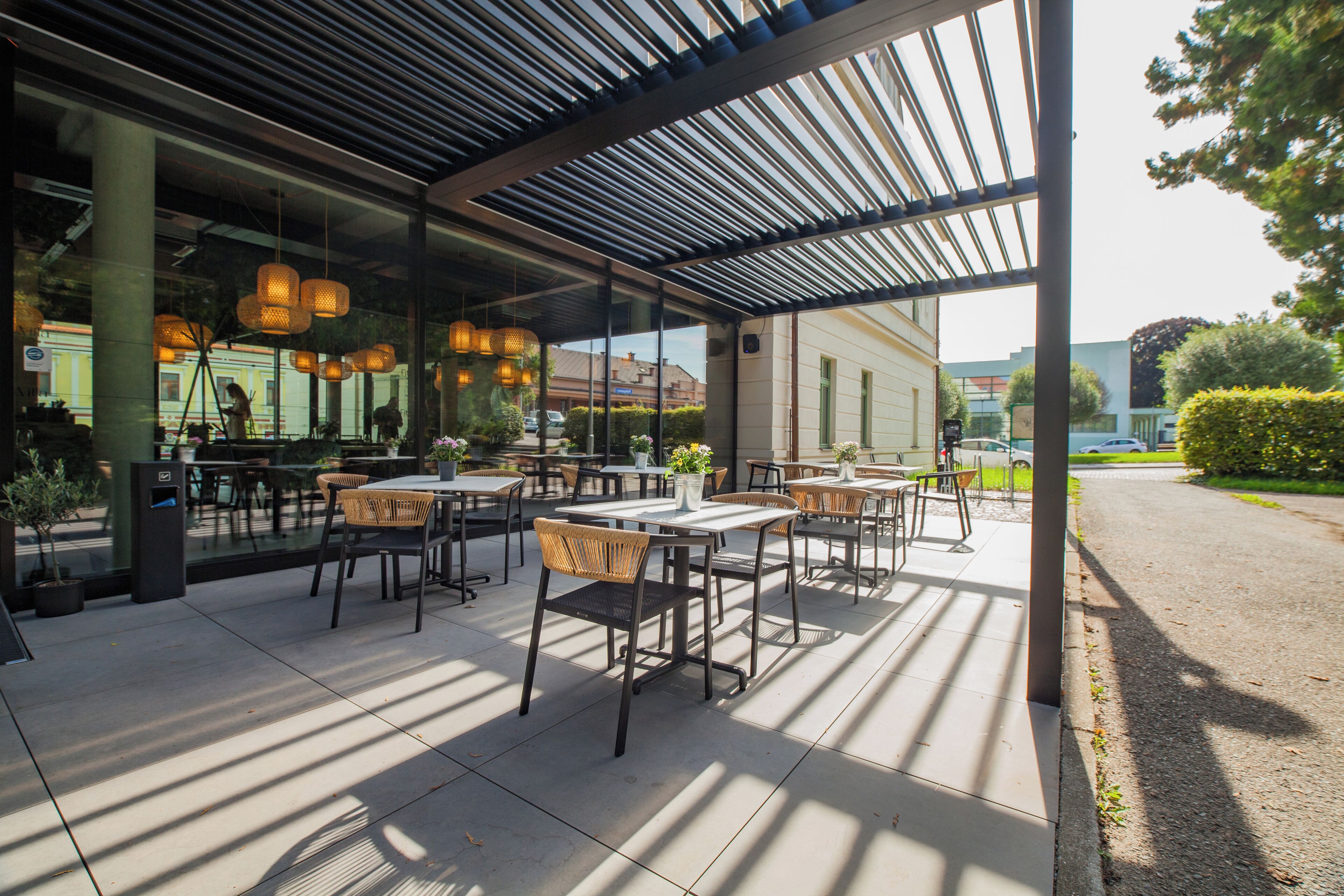 An outdoor seating area with tables and chairs, a covered section with hanging lights, and a view of the street and buildings in the background.