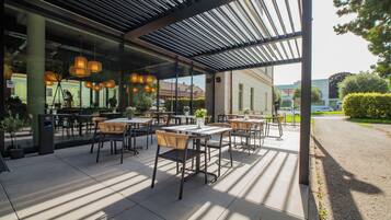 An outdoor seating area with tables and chairs, a covered section with hanging lights, and a view of the street and buildings in the background.