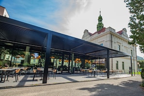 An outdoor cafe with tables and chairs, a covered seating area, and a building with a tower in the background. - Hotel Villa Fitz (Rokycany)