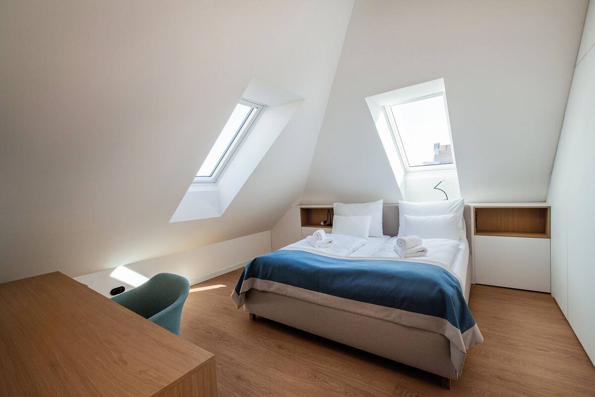 Ein modernes Schlafzimmer mit schrägem Dach, Dachfenstern, einem Bett, einem Schreibtisch und einem Stuhl.