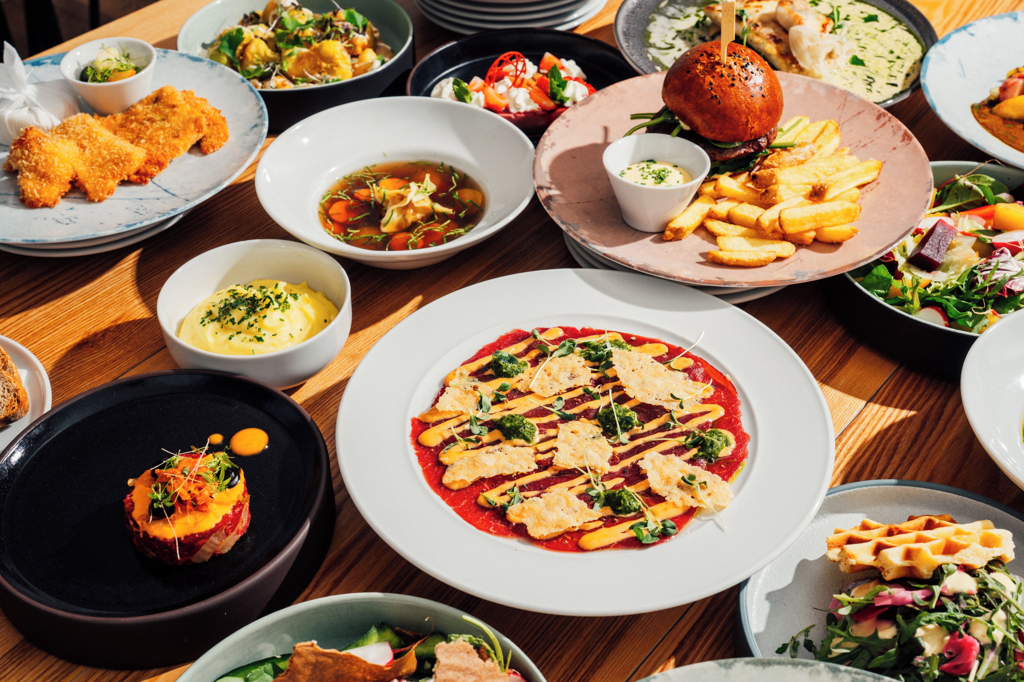 A variety of dishes on a wooden table, including a burger, fries, and a salad.