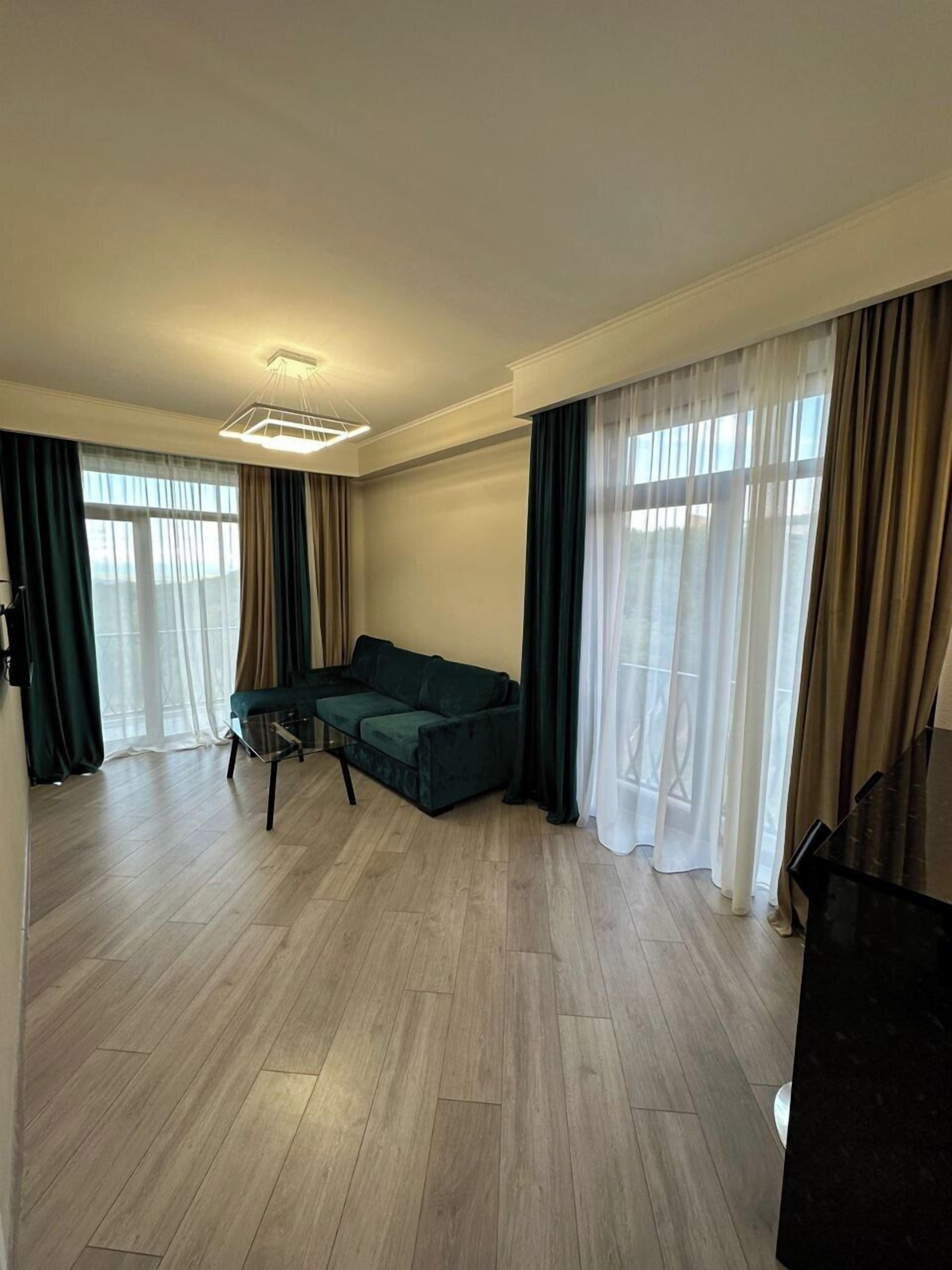 Ein geräumiges Wohnzimmer mit einem dunkelgrünen Sofa, einem Couchtisch und einem Klavier. Der Raum hat Holzböden und große Fenster mit durchsichtigen Vorhängen.