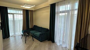 Ein geräumiges Wohnzimmer mit einem dunkelgrünen Sofa, einem Couchtisch und einem Klavier. Der Raum hat Holzböden und große Fenster mit durchsichtigen Vorhängen.