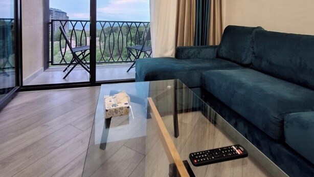 A modern living room with a dark green sofa, a glass coffee table, and a remote control on it. The room has a large sliding glass door leading to a balcony with a view of the city. - 1-3 bdr condos by Revamp (Tsaghkadzor)