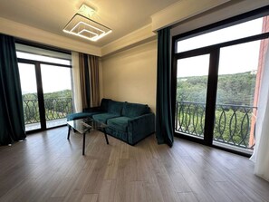 A spacious living room with a green sofa, a glass coffee table, and large windows offering a view of greenery. - 1-3 bdr condos by Revamp (Tsaghkadzor)