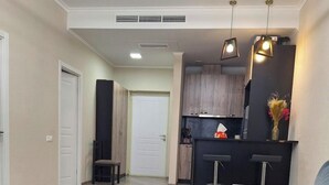 A modern living room with a glass coffee table, a grey sofa, and a kitchen area with wooden cabinets and pendant lighting. - 1-3 bdr condos by Revamp (Tsaghkadzor)