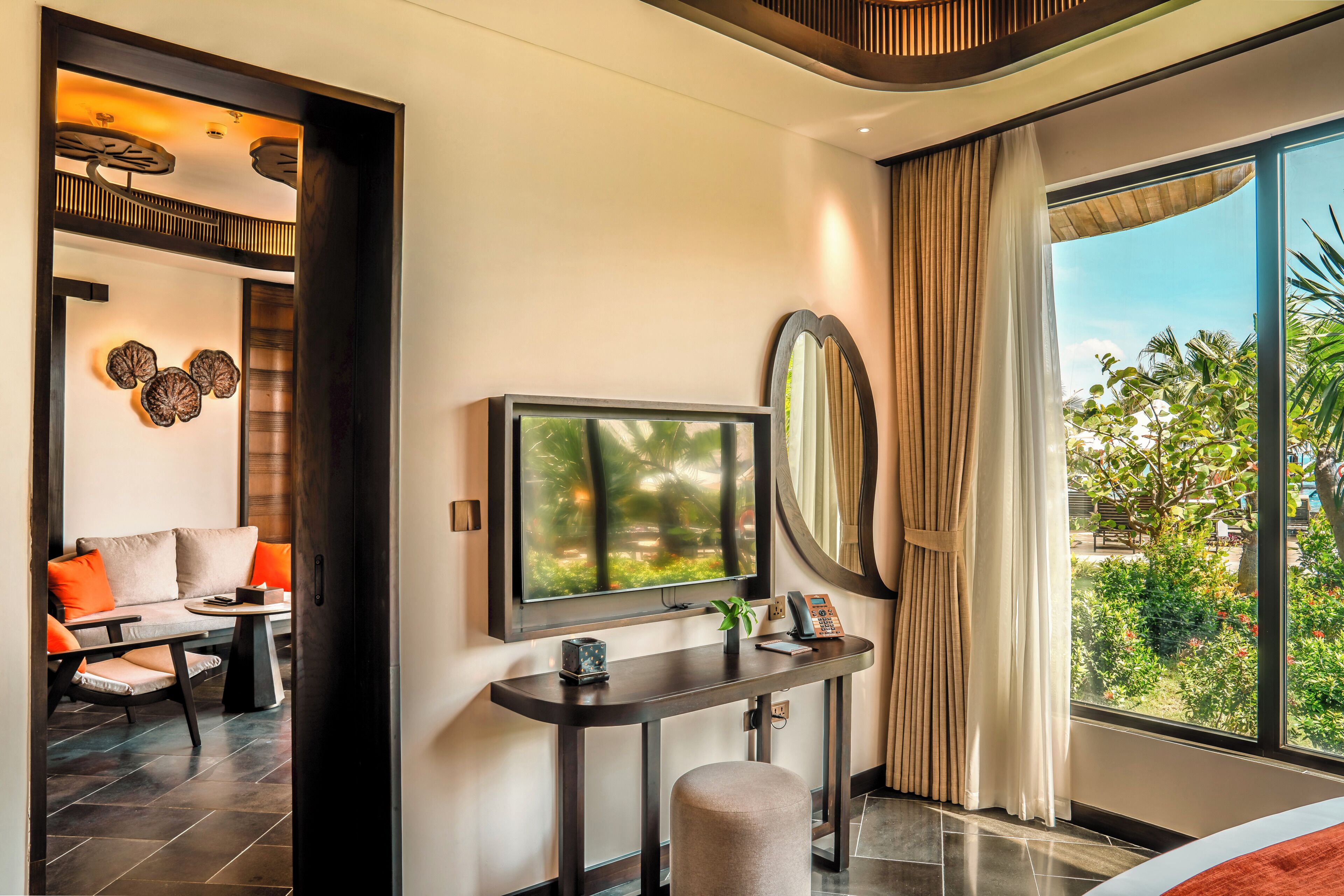 A modern hotel room with a TV, a desk, a chair, a sofa, a large window, and a view of greenery.