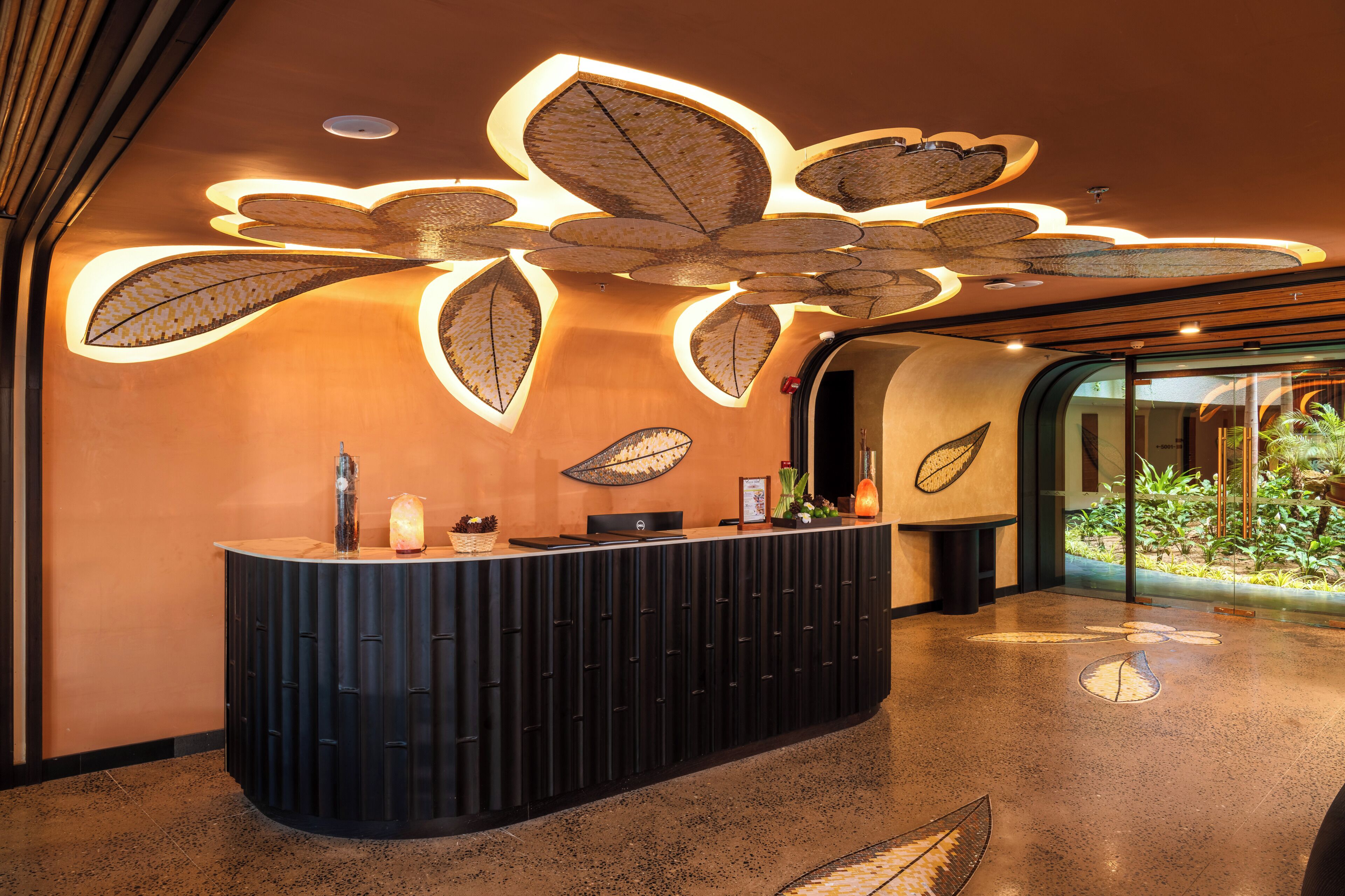 A reception desk with a modern design, a ceiling with leaf-shaped lighting, and a view of greenery through a glass door.