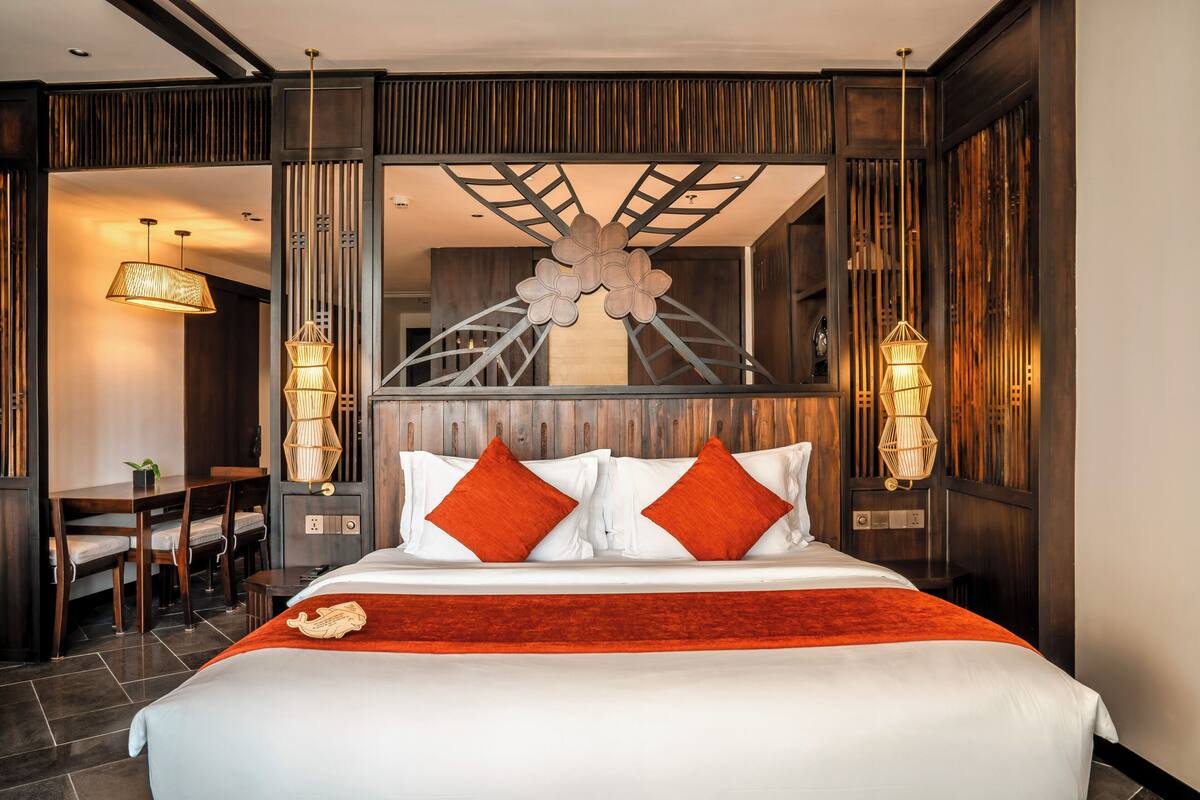 A hotel room with a large bed, two bedside lamps, a dining area with a table and chairs, and a decorative wall panel.