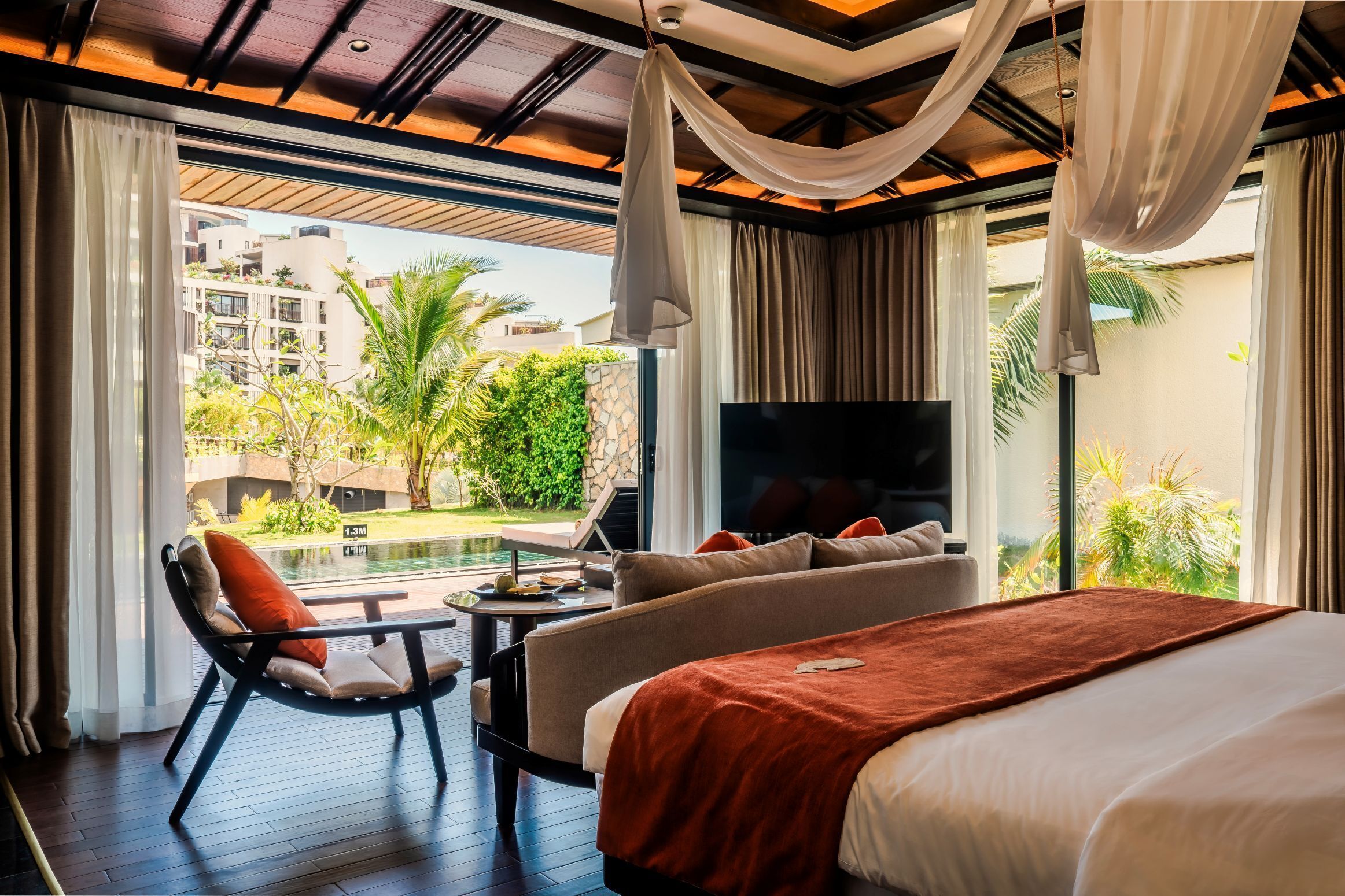A bedroom with a bed, a TV, a sofa, a chair, and a view of a pool and buildings.
