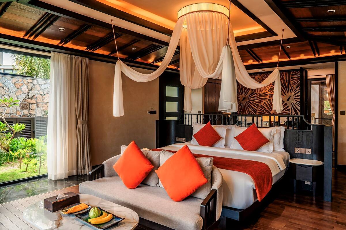 A bedroom with a bed, bedside tables, a chandelier, and a view of greenery outside.