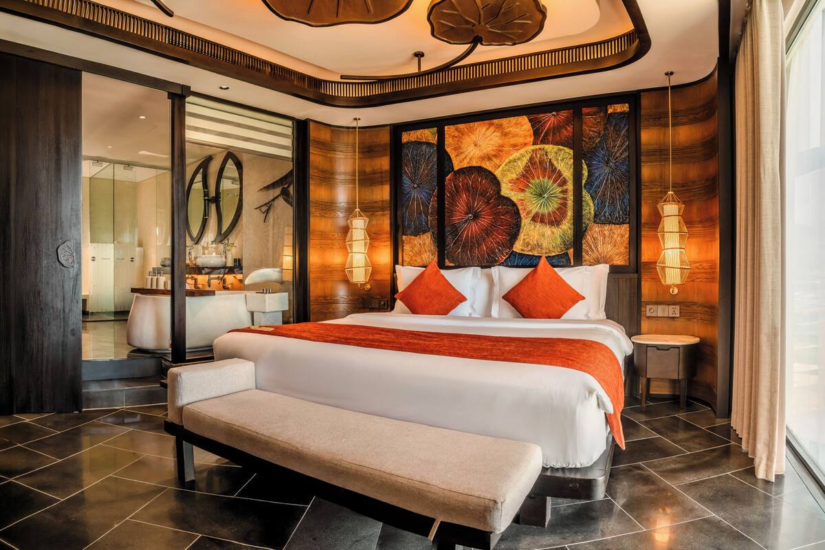 A modern hotel room with a large bed, a bench, a small table, and a large window.