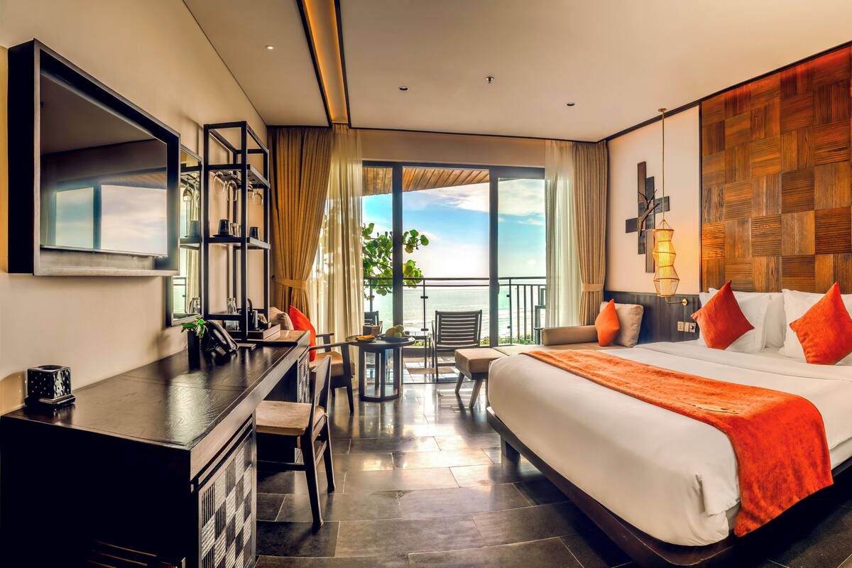 A hotel room with a large bed, a desk, a chair, a small table, and a view of the ocean.