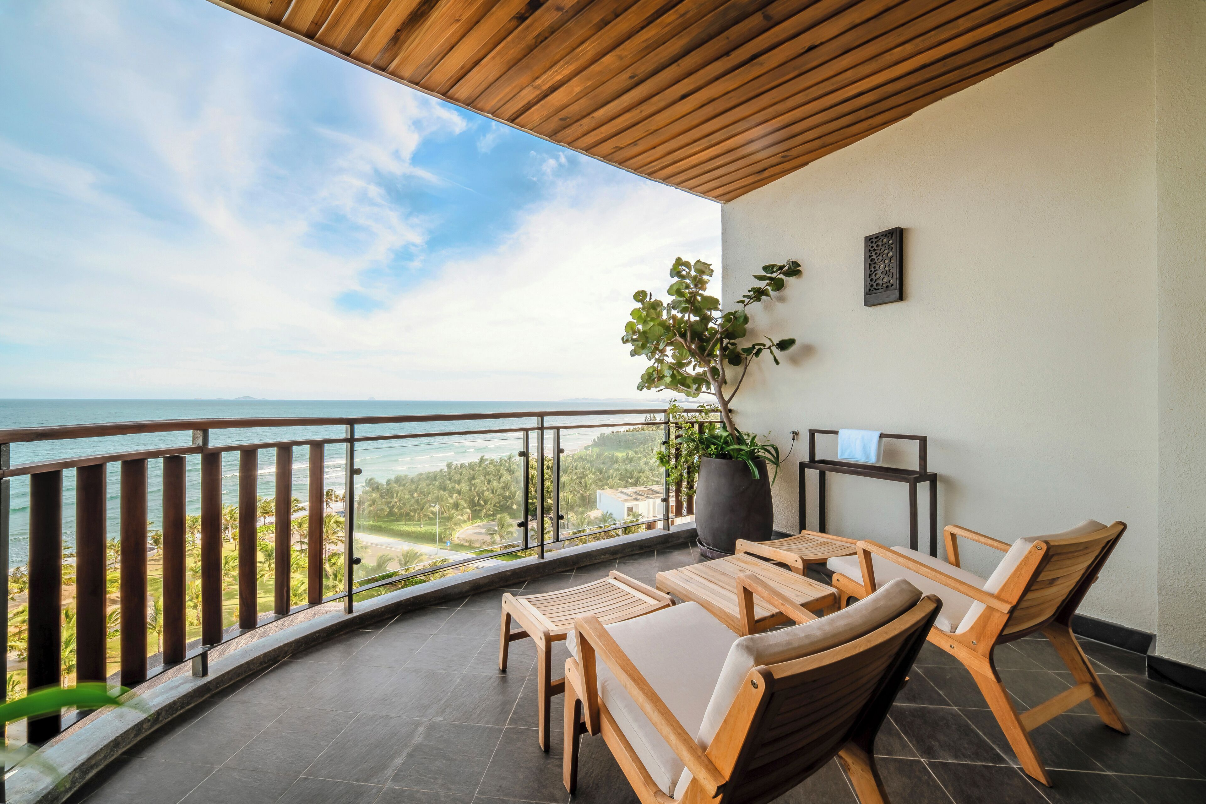 A balcony with wooden furniture, a potted plant, and a view of the sea and landscape.