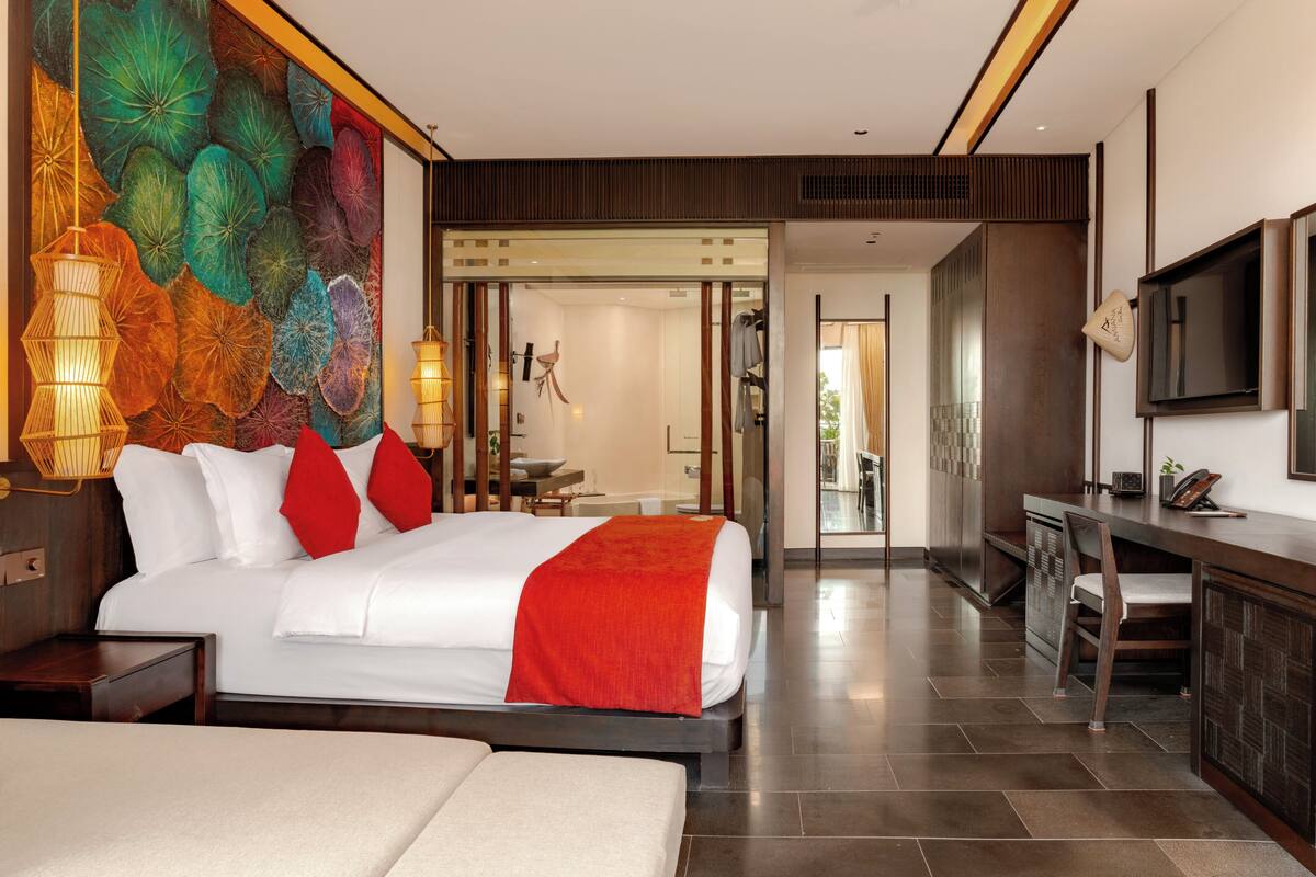 A modern hotel room with a large bed, a desk, and a view into the dining area.