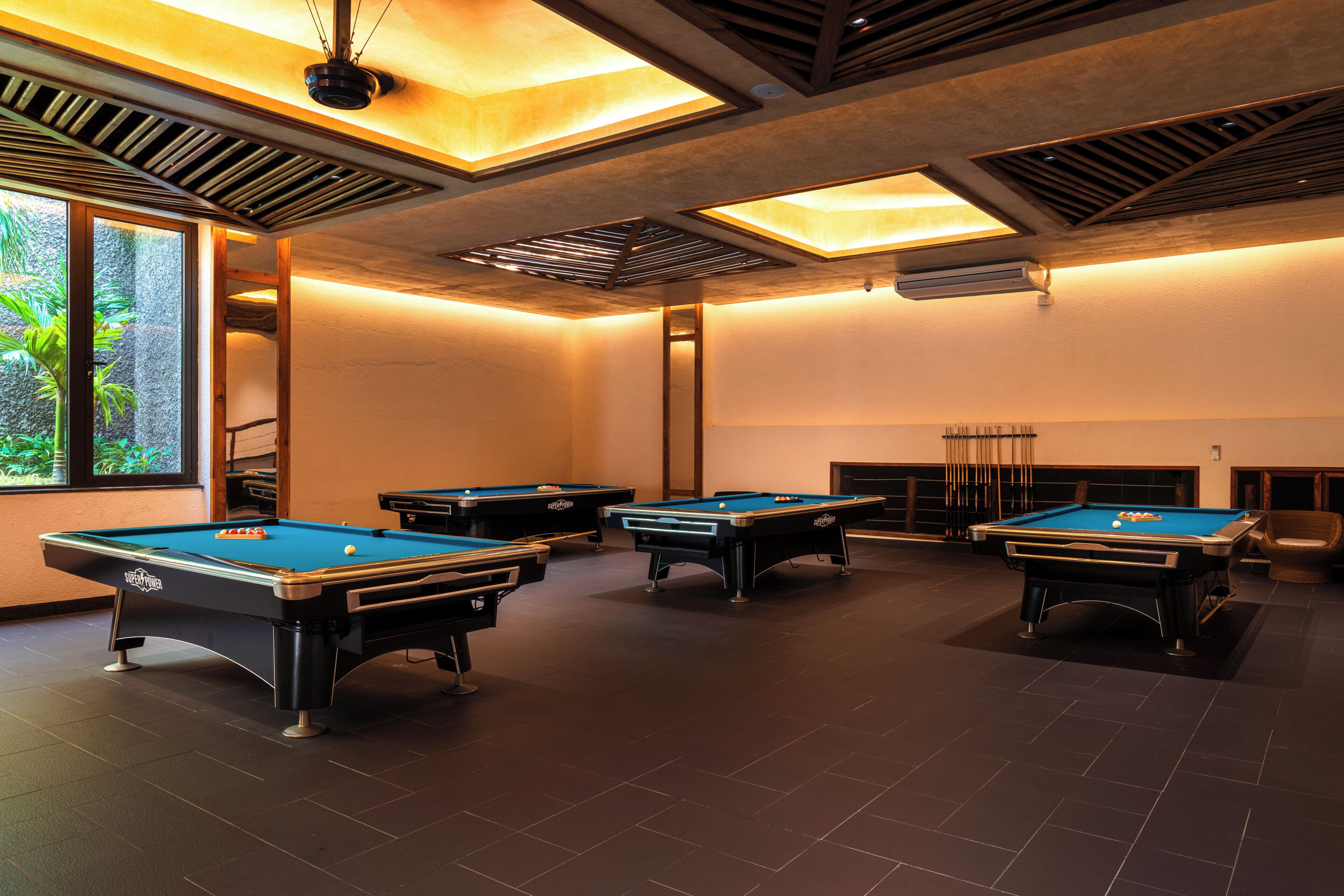 A pool room with four pool tables, a fireplace, and a large window with a view of greenery.