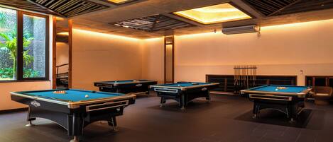 A pool room with four pool tables, a fireplace, and a large window with a view of greenery.