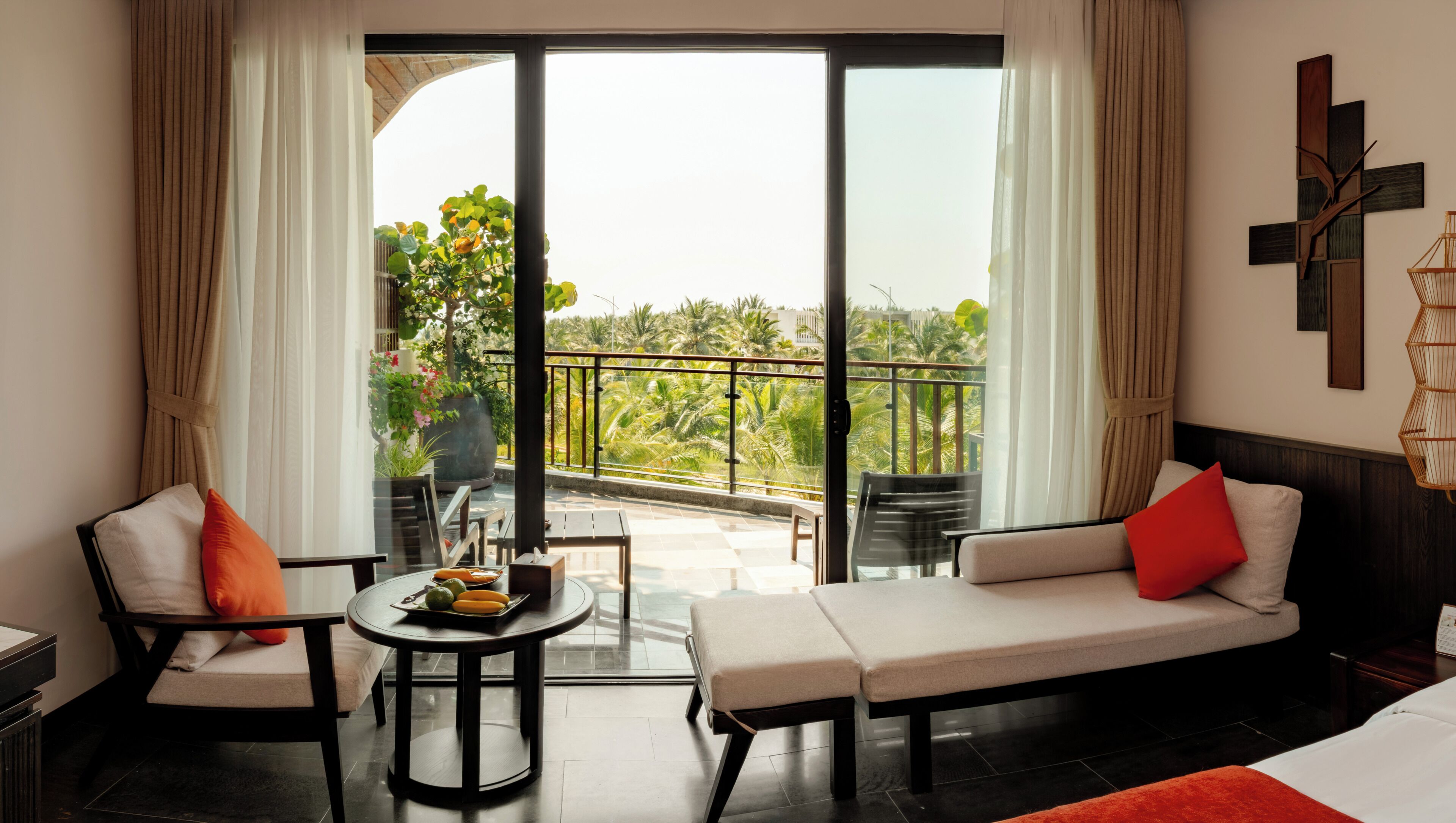 Une chambre d’hôtel avec un balcon, un canapé, une table avec des fruits et une vue sur des palmiers.